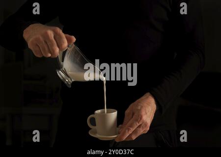 Die Hände eines nicht erkennbaren Mannes im Schatten, der Milch aus einer Glaskaraffe in eine Kaffeetasse gießt Stockfoto