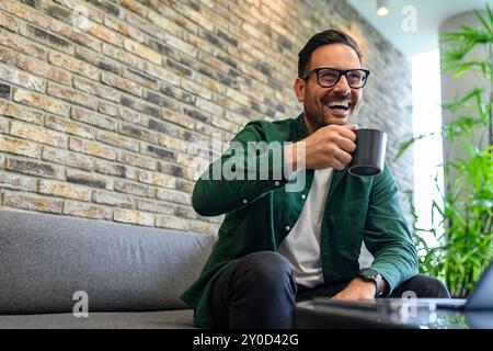 Niedriger Blickwinkel auf einen fröhlichen Geschäftsmann, der Kaffee trinkt und wegblickt, während er sich auf dem Sofa im Büro entspannt Stockfoto