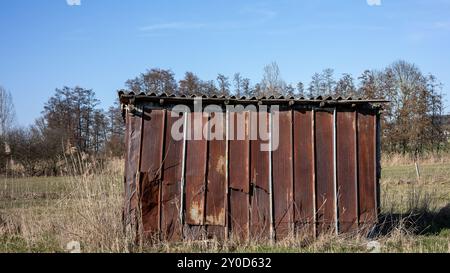 Ein Schuppen aus rostigen Metallplatten steht allein auf einem grasbewachsenen Feld, umgeben von Bäumen an einem sonnigen Tag. Stockfoto