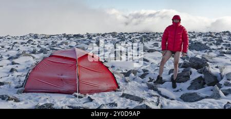 Wanderer in Hosen und Daunenjacke neben seinem Zelt auf dem Storstyggesvanatinden Berg, Dovrefjell-Sunndalsfjella Nationalpark, Oppland Fylke, Norwegen, September Stockfoto