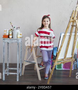 Kleine Künstler Mädchen hält einen Pinsel mit Blick auf eine Leinwand auf einer Staffelei. Konzept der Kreativität und Spaß Stockfoto