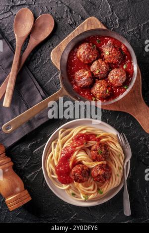 Fleischbällchen. Rindfleischbällchen, oben flach in einer Pfanne und mit einem Teller Spaghetti Nudeln, auf schwarzem Hintergrund, mit Holzlöffeln Stockfoto