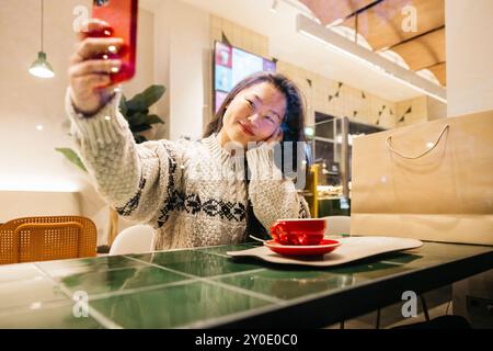 Junge asiatische Frau, die während eines Weihnachtseinkaufstages eine Pause macht. Frau, die ein Selfie in einem Café macht, mit einer Tasse Kaffee und einer Einkaufstasche neben ihr. Stockfoto