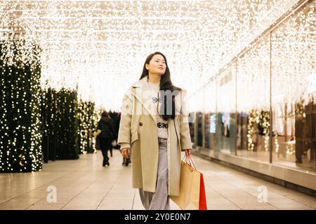 Junge asiatische Frau, die durch ein weihnachtlich dekoriertes Einkaufsviertel geht, Einkaufstaschen trägt und die festliche Weihnachtsdekoration genießt. Stockfoto