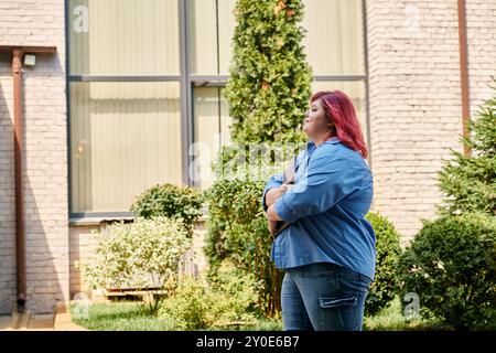 Eine Frau in Übergröße mit rotem Haar und lebhafter Kleidung schlendert durch einen sonnendurchfluteten Innenhof. Stockfoto