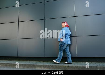 Eine kurvige Frau in Übergröße aus blauem Denim geht an einer Wand mit fließenden roten Haaren vorbei Stockfoto