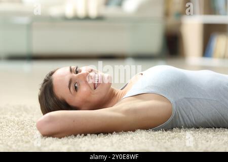 Glückliche Frau, die zu Hause auf einem Teppich auf dem Boden liegt und in die Kamera blickt Stockfoto