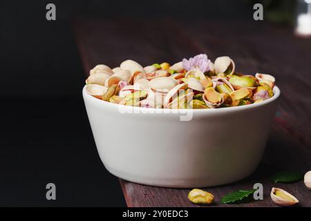 Eine weiße Schüssel gefüllt mit geschälten und ungeschälten Pistazien, auf einem rustikalen Holztisch mit dunklem Hintergrund. Stockfoto