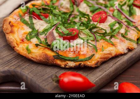 Eine köstliche neapolitanische Pizza mit Sardellen, Kapern, Kirschtomaten, frischem Gemüse und roten Zwiebeln, serviert auf einem Holzbrett Stockfoto