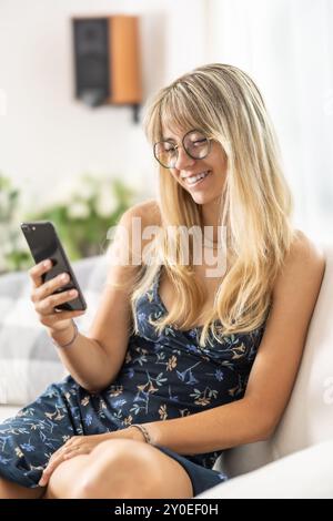 Fröhliche, entspannte junge Frau, die auf dem Sofa saß und ihr Handy benutzte, lächelnde Frau, die ihr Smartphone hielt, auf ihr Handy schaute und dabei Online-ecom spielte Stockfoto