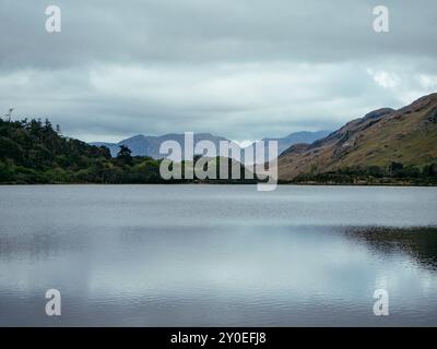 Ein ruhiger See reflektiert die grauen Wolken darüber, während üppiges Grün und zerklüftete Hügel die Szene umrahmen und eine friedliche Atmosphäre in der Natur schaffen. Stockfoto