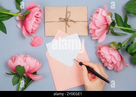 Die Hand hält einen Stift und schreibt auf leere Karte im Umschlag, rosafarbene Pfingstrosen Blumendekoration und Briefumschlag Stockfoto