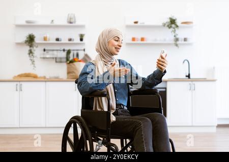 Muslimische Frau im Rollstuhl mit Smartphone in der Küche zu Hause Stockfoto