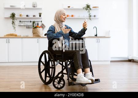 Muslimische Frau im Rollstuhl mit Handy zu Hause Stockfoto