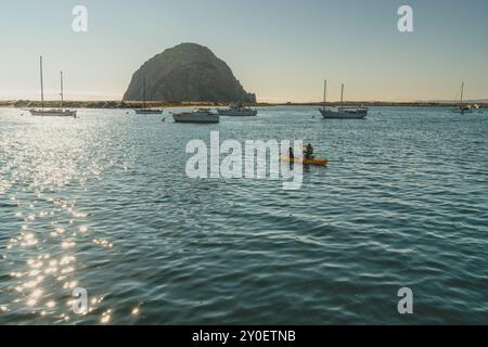 Morro Bay, Kalifornien, USA. Juli 2024. Kajakfahrer paddeln über das glitzernde Wasser mit Booten und Morro Rock im Hintergrund. Stockfoto