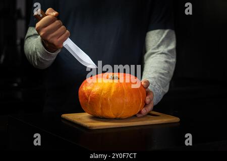 Der Halloween Herbst Orange Kürbis in Männerhand in einem schwarzen Pullover mit Küchenmesser bereitet sich auf einen Urlaub auf einem Tisch vor Stockfoto