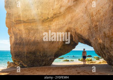 Man Tourist steht unter Klippen in Naturbogen am Sandstrand in der Nähe von Portimao in der Algarve, Portugal, Europa. Beliebtes touristisches Hotel Stockfoto