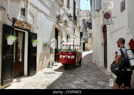 Ein rotes Tuk-Tuk-Taxi auf einer schmalen Straße; die bunten Straßen und Gassen von Monopoli puglia italien mit Touristen, Dinern, Trinken, Essen, alten Steinhäusern, Sightseeing, farbige Hutdekorationen, ein Taxi, Häuser, Balkone, Restaurants, Eis, Eis, Menschenmassen, Nacht, Tag, Melonen, Türen, Käufer. Stockfoto