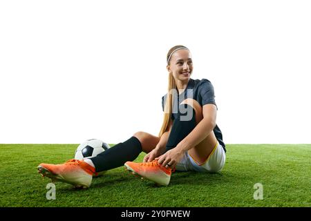 Junge Sportfrau, Mitte 20, Fußballspielerin, sitzt auf dem Feld, bindet ihre Schuhe mit Ball neben sich und lächelt, während sie sich auf die Action vorbereitet. Stockfoto