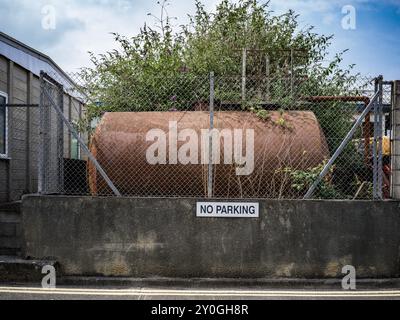 Altes, rostiges, verwittertes Ölbehältergefäß mit Umzäunung und einem Schild ohne Parken und einer niedrigen Betonwand Stockfoto