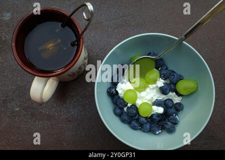 Gesundes vegetarisches Frühstück: Eine Schüssel Hüttenkäse mit Heidelbeeren und Trauben. Zusätzlich eine Tasse Tee mit einem Tee-Infuser Stockfoto