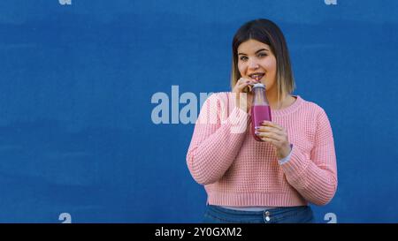 Lächelnde junge Frau mit Ombre-Haar, die einen rosa Smoothie in einer Glasflasche genießt. Sie trägt einen rosafarbenen Strickpullover vor einem leuchtend blauen Hintergrund Stockfoto