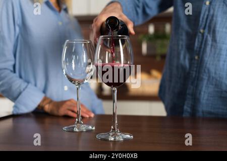 Nicht erkennbarer Mann, der einen Rotwein in ein Glas in Blasen mit einem Spritzer auf dem Tisch in der Küche eingoß, Nahaufnahme Stockfoto