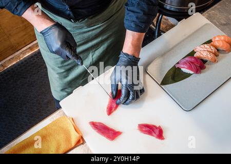 Nahaufnahme des Küchenchefs mit schwarzen Handschuhen, der rohen Thunfisch für Sushi schneidet. Professioneller Koch bereitet frischen Fisch auf Schneidebrett mit Nigiri-Sushi im Backgro zu Stockfoto