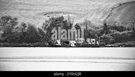 Holzboote mit Masten und Seilen sind an einem Sandstrand geerdet. Das Boot ist verwittert und in Verfall. Im Hintergrund ist gepflügtes Land. Stockfoto