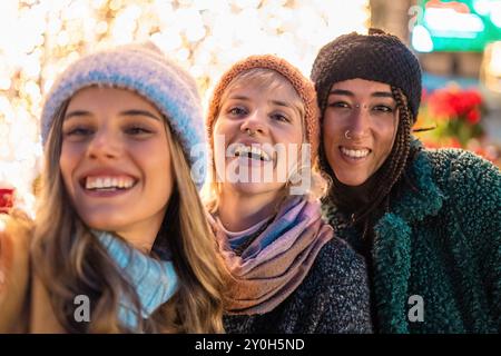 Nahaufnahme von drei verschiedenen jungen Freundinnen, die lachen und gemeinsam die Feiertage genießen. Trendige Wintermode mit festlichen Lichtern hinten Stockfoto