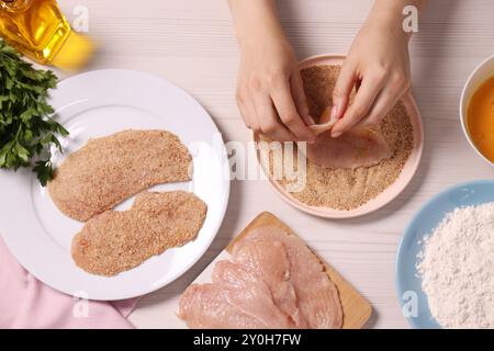 Schnitzel machen. Frau, die Fleischscheiben mit Brotkrümeln an weißem Holztisch überstrich, Blick von oben Stockfoto