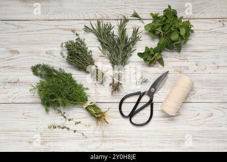 Bündel von verschiedenen frischen Kräutern, Spule mit Faden und Schere auf weißem Holztisch, flach gelegt Stockfoto