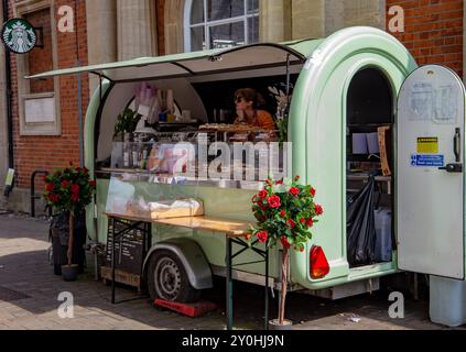 Ein Food Truck im Vintage-Stil, der eine Auswahl an Gebäck, Kaffee und Getränken anbietet, parkt vor einem alten Backsteinhaus. Stockfoto