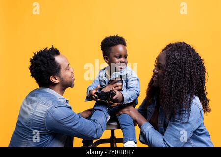 Stolze junge Eltern spielen mit ihrem niedlichen Sohn im Studio und umarmen ihn vor gelbem Hintergrund. afroamerikaner-Mutter und Vater posieren mit ihrem kleinen Kind, das einen Kontrolleur hält. Stockfoto