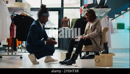 Der Verkäufer empfiehlt ein Paar Stiefel für schwangere Frauen und hilft Ihnen dabei, die perfekte Größe zu finden. afroamerikanischer Kunde probiert Schuhe im Bekleidungsgeschäft an, Konsum. Kamera B.. Stockfoto
