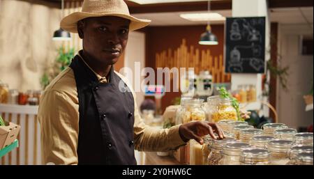 afroamerikanischer Verkäufer, der lokale Bauern vermarktet und Waren in wiederverwendbaren Gläsern auf der Ladenpräsentation anordnet. Junger Erwachsener, der Waren in einem Supermarkt in der Nachbarschaft verkauft. Kamera 2. Stockfoto