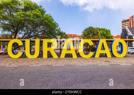 Malerische Aussicht auf große gelbe „Curacao“-Buchstaben auf dem Marktplatz im Stadtzentrum, mit Gebäuden, blauem Himmel und Bäumen im Hintergrund. Willemstad. Curacao. Stockfoto