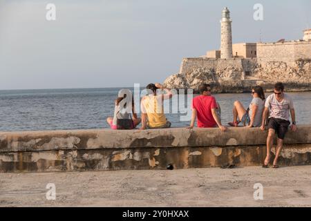 Kuba, Havanna, Junge Leute sitzen auf der Ufermauer in der Nähe des Malecon. Morro Festung im Hintergrund. 24. März 2016 Stockfoto