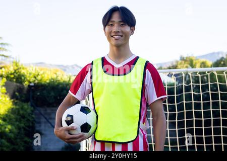 Fußball halten, junger asiatischer Mann in Sportuniform lächelt durch Tornetz Stockfoto