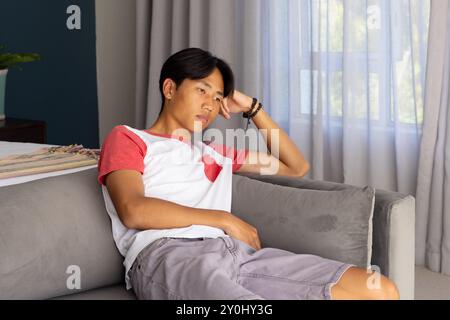 Entspannend auf der Couch, junger asiatischer Mann in lässiger Kleidung, der nachdenklich aussieht Stockfoto