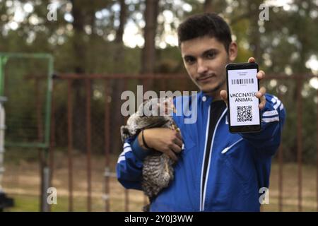 Porträt eines jungen Landwirts, der auf seinem Handy den Impfpass seiner Hühner zeigt, während er eine Hühnerin in den Armen hält. Tierimpftasse Stockfoto