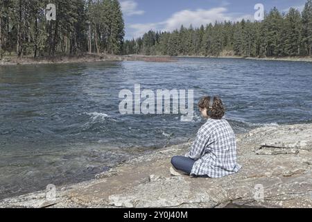 Eine junge Frau sitzt am Spokane River in Post Falls, Idaho, entspannt an einem sonnigen Tag und hört Musik über ihre Kopfhörer Stockfoto