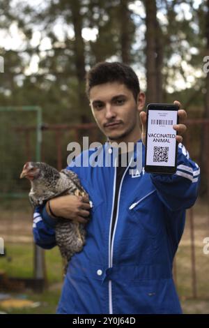 Vertikales Porträt eines jungen Landwirts, der auf seinem Handy den Impfpass seiner Hühner zeigt, während er eine Hühnerin in den Armen hält. Tierimpfstoff Stockfoto