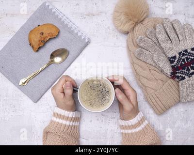 Ein flaches Foto von den Händen einer Frau, die eine Tasse Kaffee, einen Holzlöffel, Kekse, Handschuhe und einen Hut an der Seite hält Stockfoto