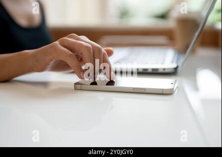 Ein Nahbild eines weiblichen Fingers, der auf einem Tisch einen Smartphone-Bildschirm berührt. Eine Frau, die aus der Ferne von einem Café aus arbeitet und ihr Smartphone benutzt Stockfoto