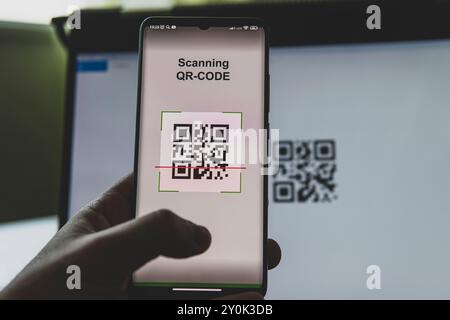 Junger Geschäftsmann, der an seinem Arbeitsplatz vor einem Computermonitor sitzt und QR-Code mit dem Handy scannt. Stockfoto