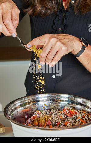 Nahaufnahme einer Person, die gemahlene Gewürze über einen bunten Salat in einer Edelstahlschüssel in der Küche streut. Stockfoto