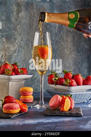 Champagner wird in eine Flöte mit einer Erdbeere gegossen, umgeben von frischen Erdbeeren und bunten Macarons auf einer strukturierten Oberfläche. Stockfoto