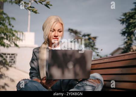 Eine blonde Frau sitzt auf einer Bank mit einem Laptop vor sich. Sie trägt eine graue Jacke und Jeans. Die Szene deutet auf eine lässige und entspannte Atmosphäre hin Stockfoto