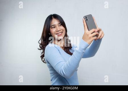 Schöne langhaarige asiatische Frau macht Selfie mit ihrem Handy in blauem Strickoberteil, weißer Hintergrund Damenbluse Stockfoto